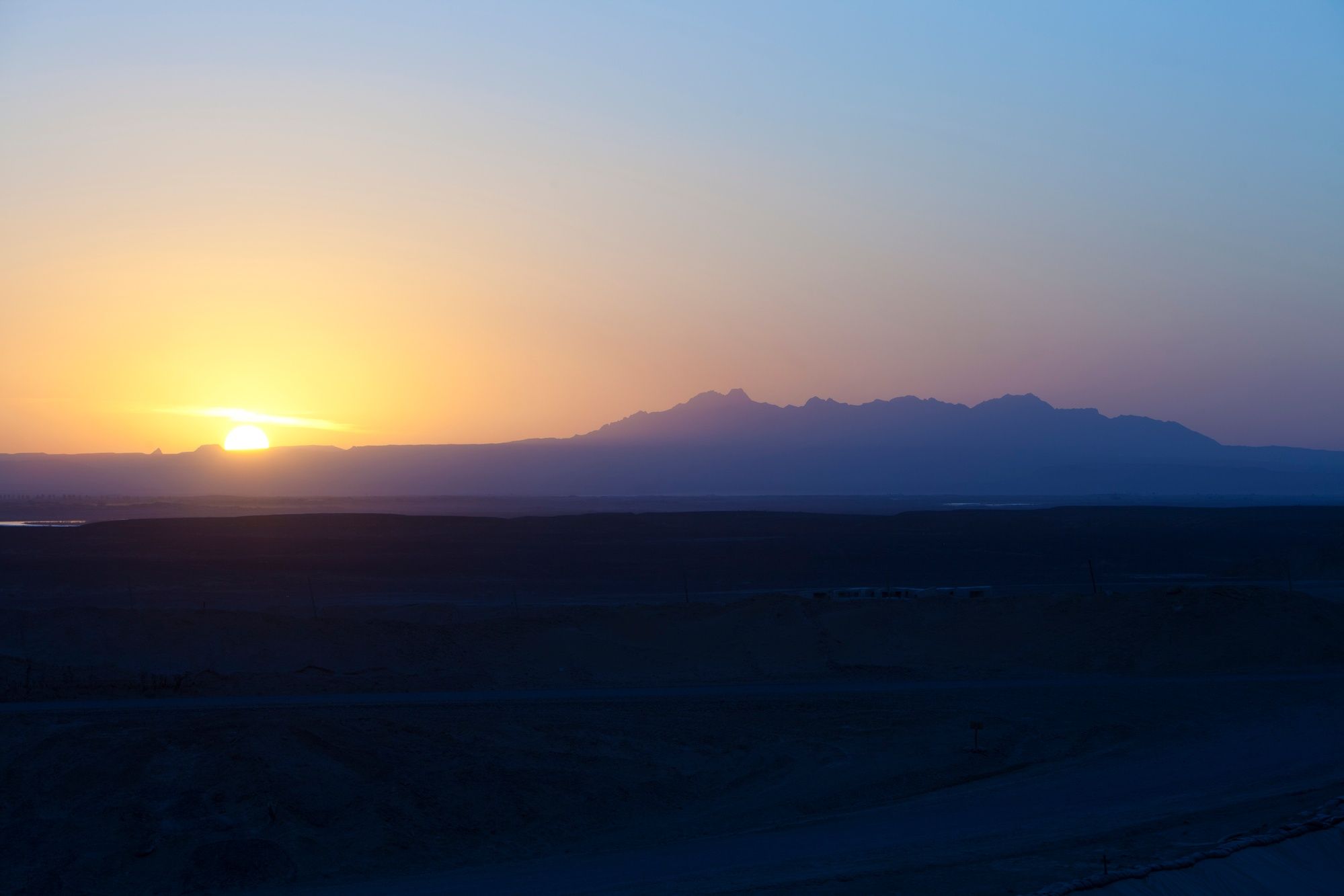 The sun sets behind a line of mountains over a darkening desert plain.
